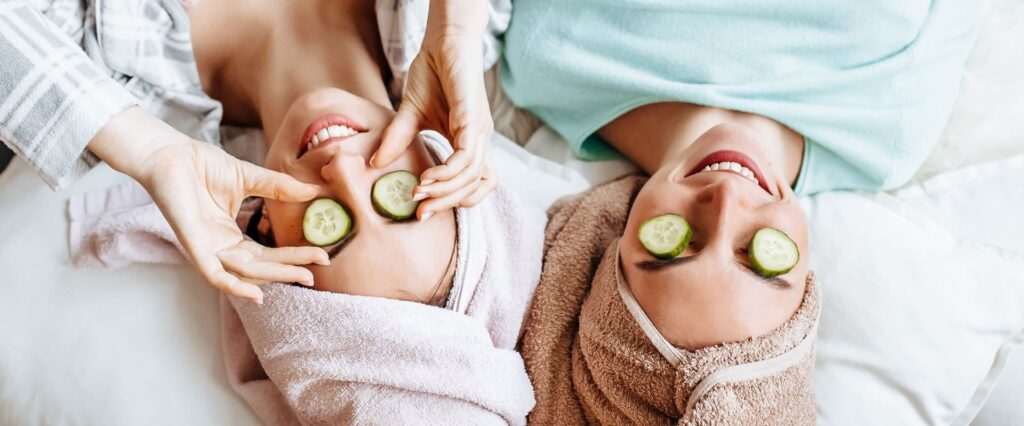 Zwei Frauen liegen entspannt mit Gurkenscheiben auf den Augen und Handtüchern um die Haare gewickelt. Sie genießen eine Wellness-Behandlung zu Hause, während moderne Badarmaturen im Badezimmer für ein luxuriöses Ambiente sorgen.