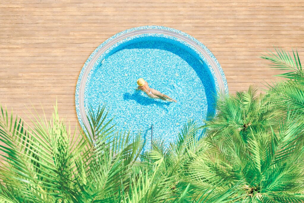 Runder Swimmingpool mit Holzumrandung und tropischen Pflanzen aus der Vogelperspektive